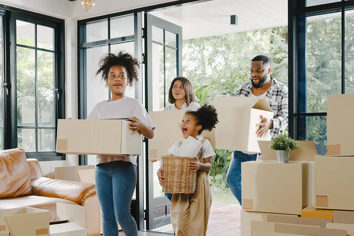 A family of four - father, mother, and 2 daughters - entering their new house carrying boxes.